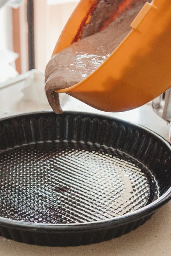 Chocolate cake batter being poured from an orange jug into a dark fluted baking tin