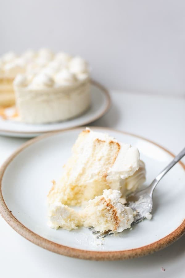 A single slice of layered white cake on a plate with a fork, the rest of the cake on a stand behind it