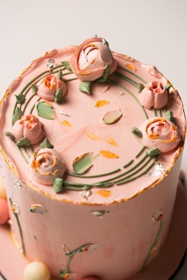 A tall cake finished in pale pink buttercream, its top edge piped with buttercream roses and green leaves