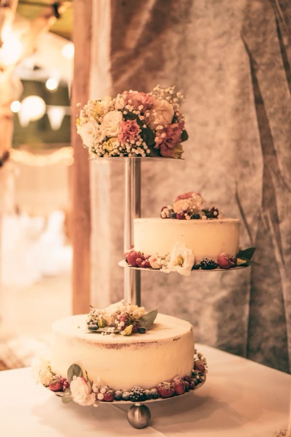 Three separate cake tiers on a tall metal stand, each finished in pale buttercream and topped with flowers and berries