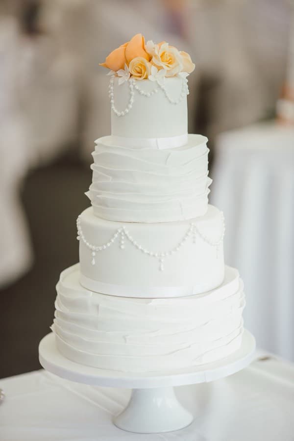 A four-tier white wedding cake stacked on a pedestal stand, with ruffled fondant tiers and piped pearl swags
